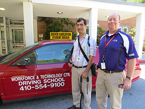 Two men stand next to a car. On the side of the car are the words Workforce & Technology Center Driving School 410-554-9100. A sign on the roof says Driver Education Student Driver.